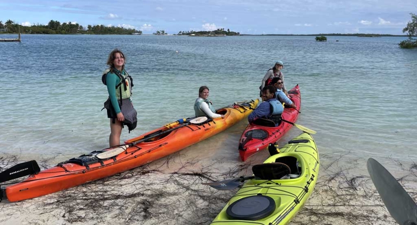 People gather beside colorful kayaks beached near clear blue water